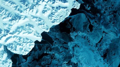 Aerial view of blue ice and water. A volcanic terrain is hidden under snow-covered peaks and valley glaciers feed blue ice into coastal waters